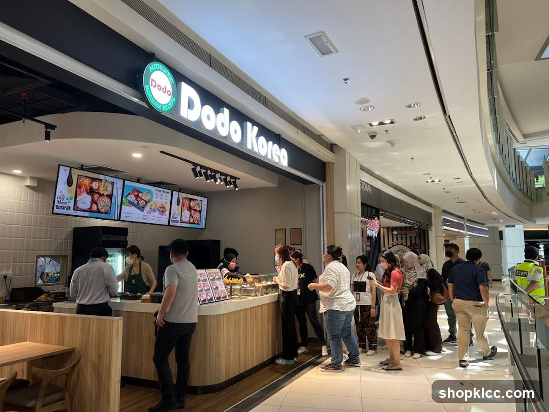 A line of customers waits to order at a Dodo Korea food stall. The stall has a prominent sign with the Dodo Korea logo and name. Digital menus displaying food items are mounted above the counter. Staff members are visible behind the counter, serving customers. The stall is located in a mall or shopping center, with other shops visible in the background.