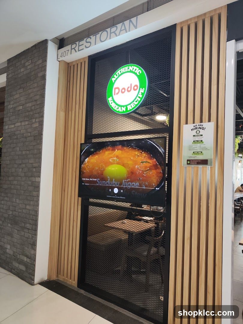 The exterior of a restaurant with a sign that says "RESTORAN" and a circular green sign with "AUTHENTIC Dodo KOREAN RECIPE" written on it. A screen displays a video of a dish called "Sundubu Jjigae Spicy Soft Tofu Stew". There is also a "WE ARE HIRING!" poster on the right side.