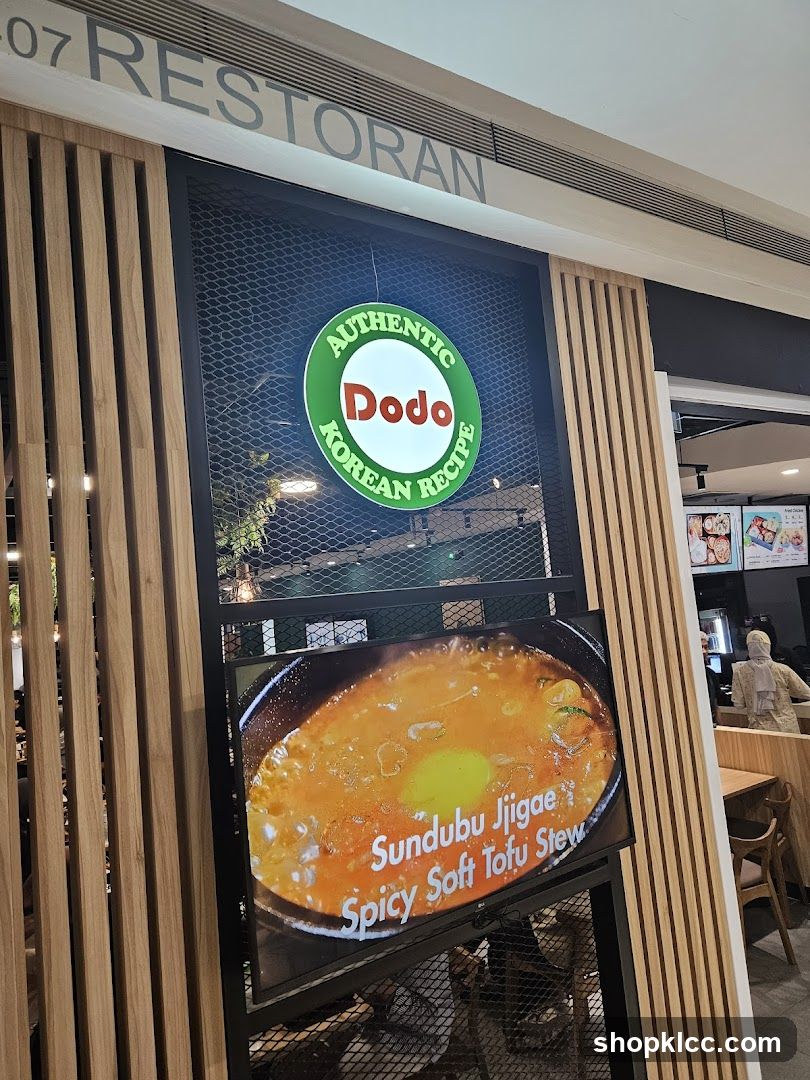 The exterior of a restaurant with a sign that says "RESTORAN" above a circular green and white sign with "AUTHENTIC Dodo KOREAN RECIPE" written on it. Below the sign is a digital display showing a close-up of Sundubu Jjigae, described as "Spicy Soft Tofu Stew". The restaurant has wooden slat paneling on the facade.