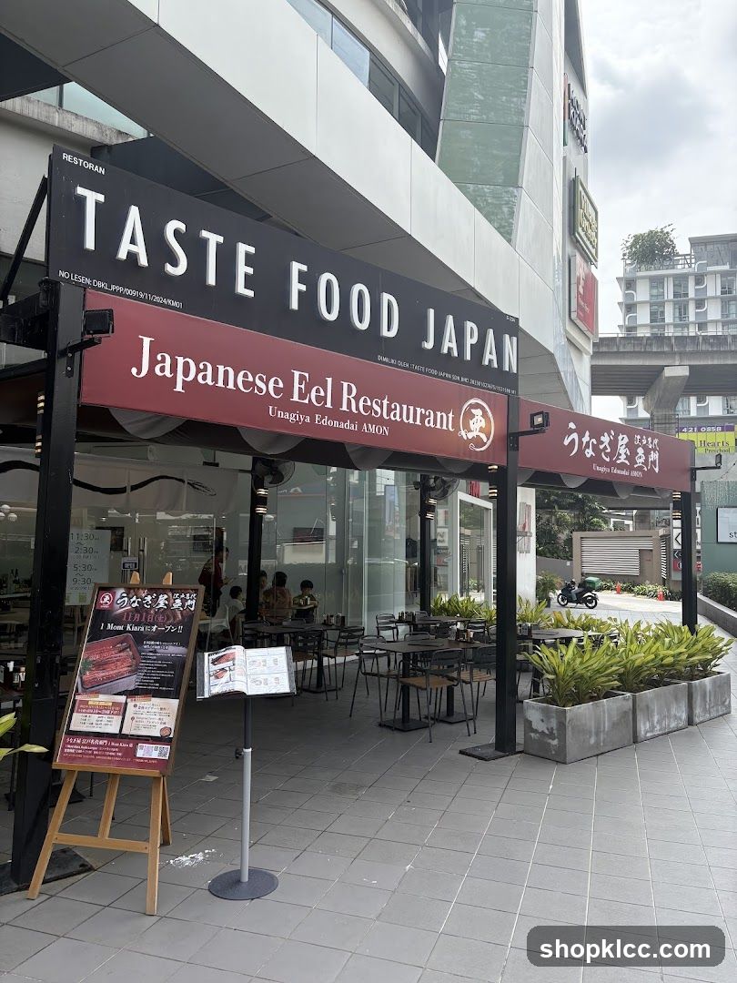 The exterior of a Japanese eel restaurant with outdoor seating. A large sign above the entrance reads "TASTE FOOD JAPAN" and "Japanese Eel Restaurant". Below the sign, there is an easel displaying a menu with images of eel dishes and Japanese text.