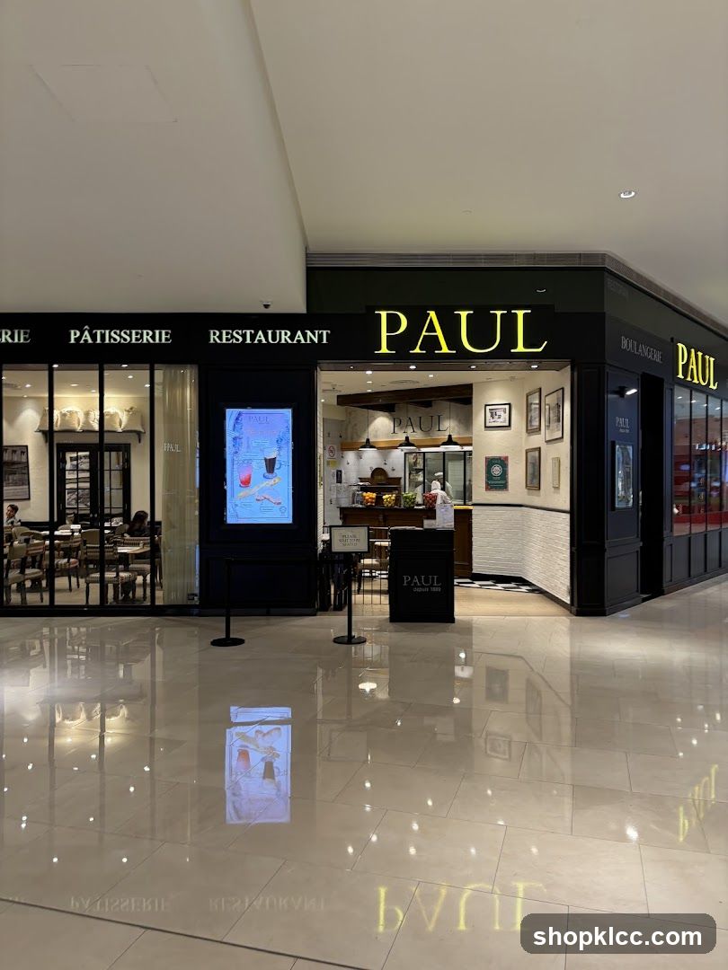 The exterior of a PAUL boulangerie, patisserie, and restaurant. The storefront features large windows displaying seating areas and food items. Prominent signage with the PAUL logo is visible above the entrance and on the building facade.