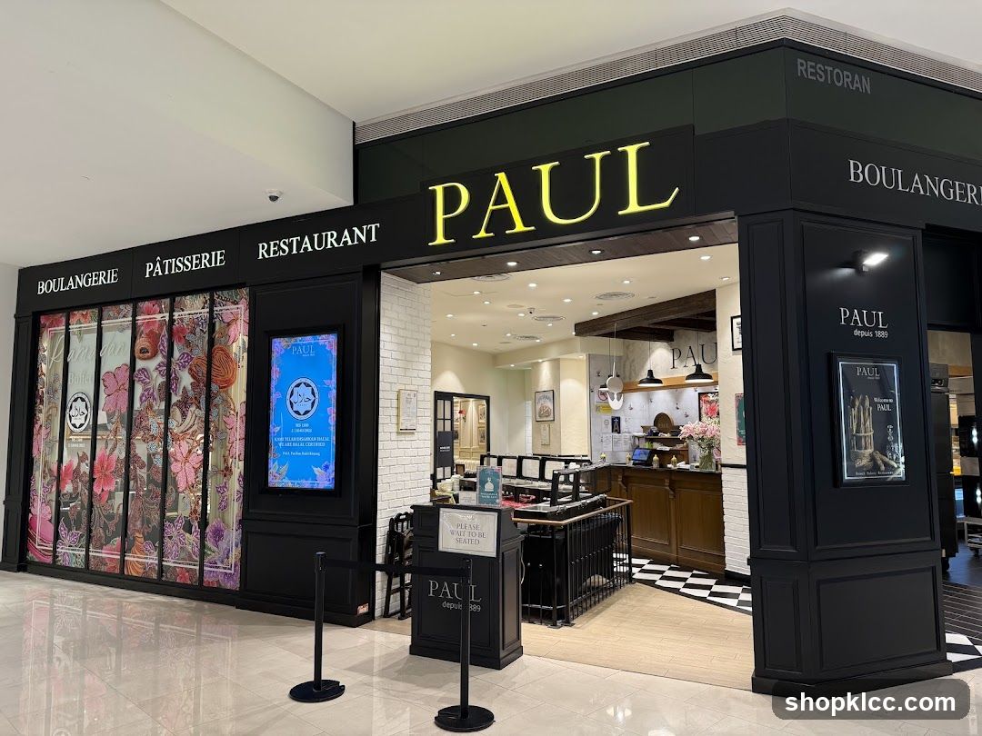 The exterior of a Paul Boulangerie Pâtisserie Restaurant. The storefront features large windows with decorative floral patterns and signage for "PAUL" in large yellow letters above the entrance. To the left, "BOULANGERIE PÂTISSERIE RESTAURANT" is visible. To the right, "RESTORAN BOULANGERIE" is displayed. A digital screen shows a halal certification. A framed poster with "PAUL depuis 1889" is also visible on the right side.