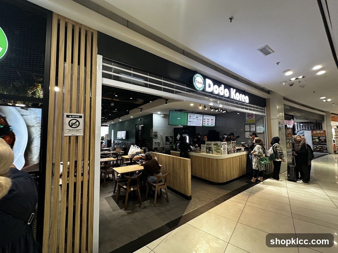 The exterior of a Dodo Korea restaurant with a prominent sign above the entrance. Inside, customers are seen ordering and dining at tables. The restaurant has a modern interior with wooden accents.
