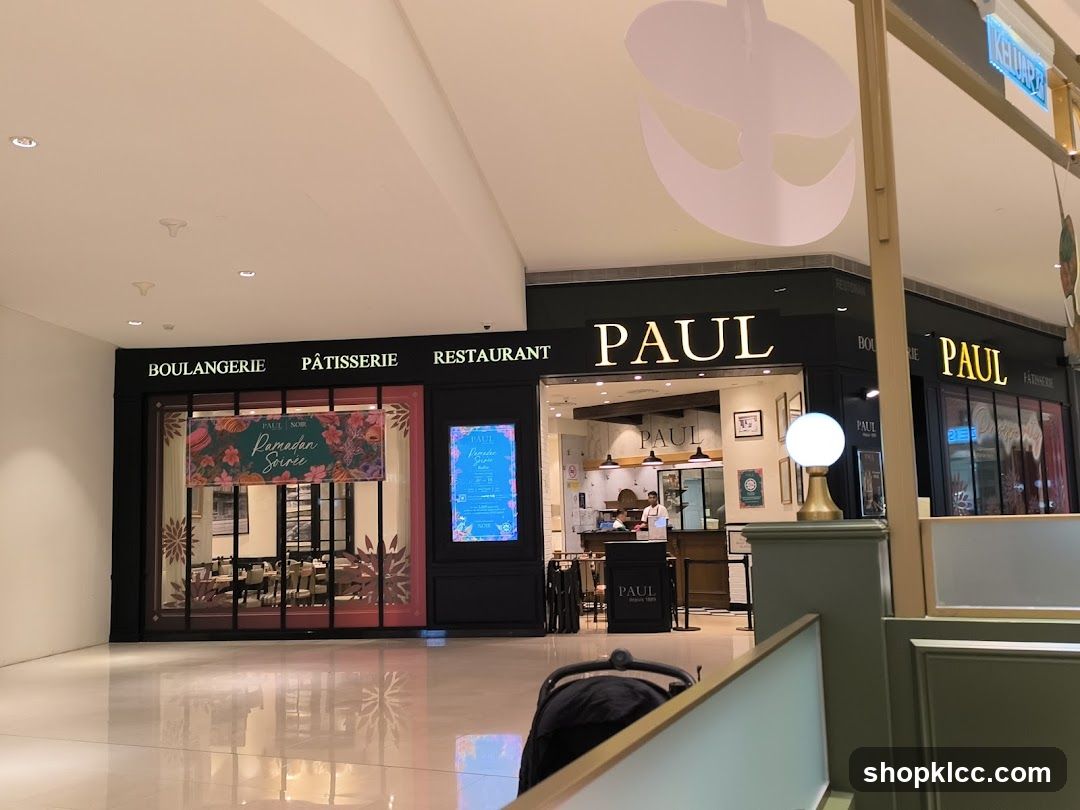 The exterior of a PAUL Boulangerie Pâtisserie Restaurant. The storefront features large windows and prominent signage with the PAUL logo. Inside, tables and chairs are visible, set up for dining. A banner advertising "Ramadan Soirée" is displayed in one window.