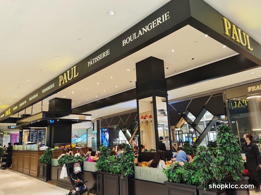 The exterior of a PAUL Boulangerie Pâtisserie restaurant with outdoor seating. The restaurant has a prominent sign with the name "PAUL" in gold lettering, along with "BOULANGERIE" and "PÂTISSERIE". There are tables and chairs set up outside, surrounded by greenery. People are seated at the tables, and staff members are visible. The interior of the restaurant is visible through the windows, showing more seating and displays.