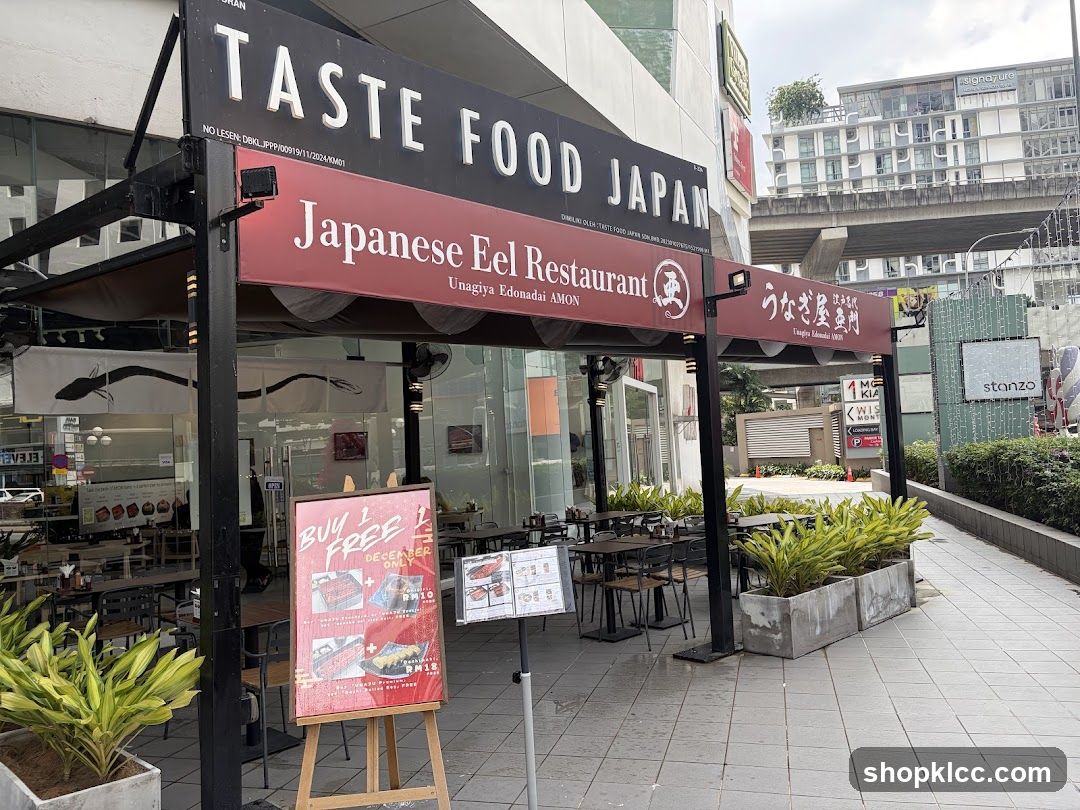 The exterior of a Japanese eel restaurant with outdoor seating. The restaurant has a large black sign with white lettering that reads "TASTE FOOD JAPAN" and a red banner below that says "Japanese Eel Restaurant" and "Unagiya Edonadai AMON". There are tables and chairs set up outside, and potted plants in front of the building. A promotional poster is displayed on an easel.