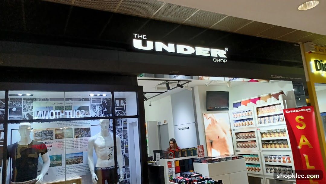 The exterior of a retail store called "The Under Shop". The store has a large black sign with illuminated white text that reads "THE UNDER SHOP". The storefront features mannequins displaying clothing and shelves stocked with merchandise. A red banner with "UNDER SALE" is visible on the right side.