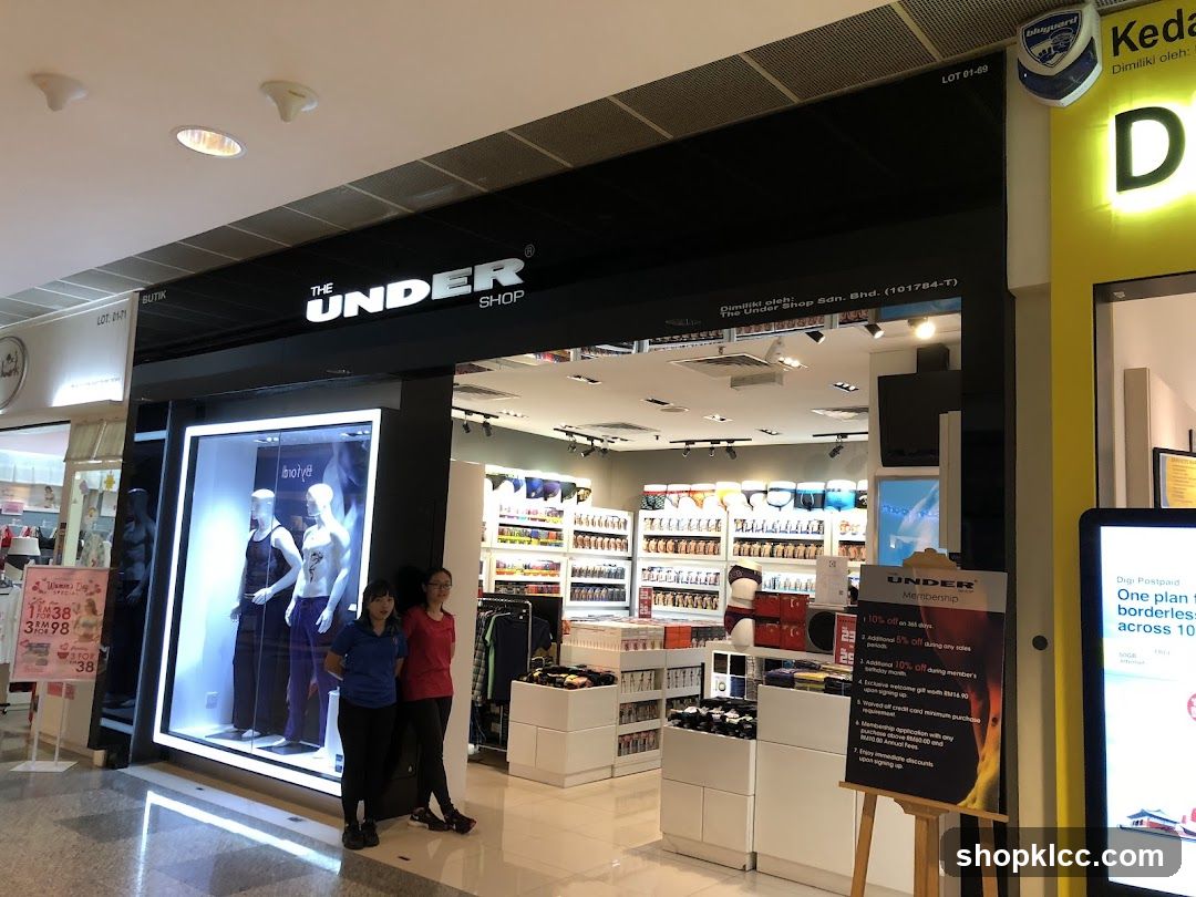 The exterior of a retail store called The Under Shop. The store has a large display window with mannequins wearing underwear. Inside, shelves are stocked with various undergarments. Two employees stand near the entrance. A promotional sign for membership is displayed outside the store.