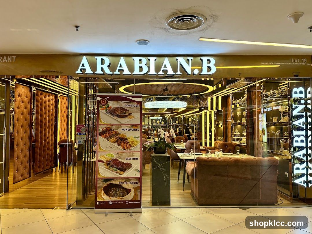 The exterior of a restaurant with a large illuminated sign that reads "ARABIAN.B". The entrance features glass doors and windows, revealing a well-lit interior with plush seating and decorative elements. A vertical banner displays images of various dishes, including Lamb Ribs, Mixed Grill, Beef Ribs, and Kofta Tahini.