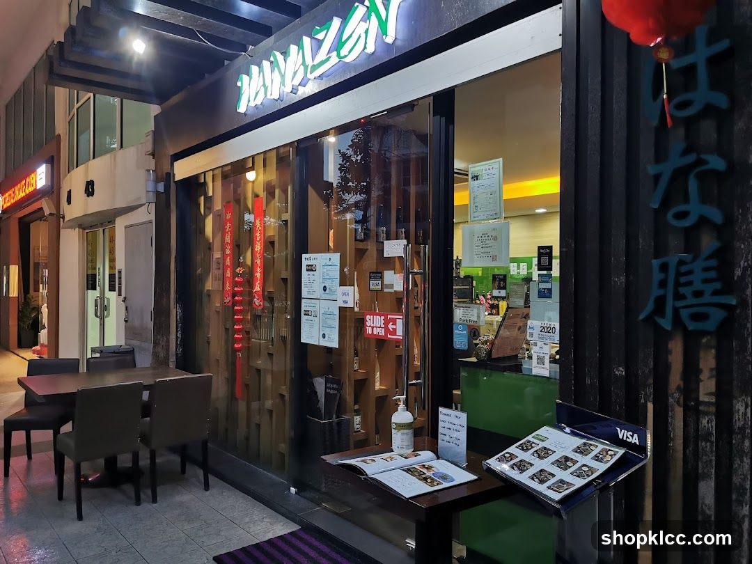 The exterior of a Japanese restaurant with a prominent sign that reads "HAYAZEN" in illuminated letters. The storefront features large glass windows and wooden paneling. Outside, there are tables and chairs set up for dining. A menu stand is placed on a table near the entrance, displaying various dishes.