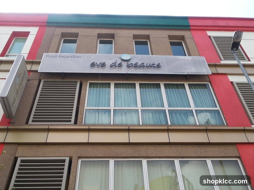 A close-up, low-angle shot of the exterior of a building with a sign for "Eve de Beaute" and "Pusat Kecantikan". The building has a modern facade with multiple windows and red and green accents on the upper levels. A street lamp is visible on the right side.