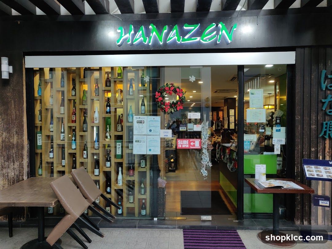 The exterior of a Japanese restaurant called Hanazen. The storefront features a large glass window displaying numerous bottles of sake on shelves. A wreath hangs on the door, and tables and chairs are set up outside. The restaurant's name is illuminated in green neon above the entrance.