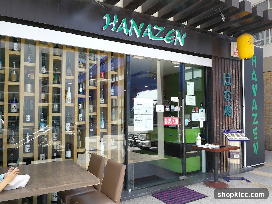 The exterior of a Japanese restaurant called Hanazen. The storefront features large glass windows displaying shelves filled with bottles, likely sake. A prominent green sign with the restaurant's name is visible above the entrance. Vertical signage with Japanese characters and the name "HANAZEN" is also present on the right side of the entrance. A yellow lantern hangs near the entrance. Outside, a table and chairs are set up, and a menu stand is placed near the door.