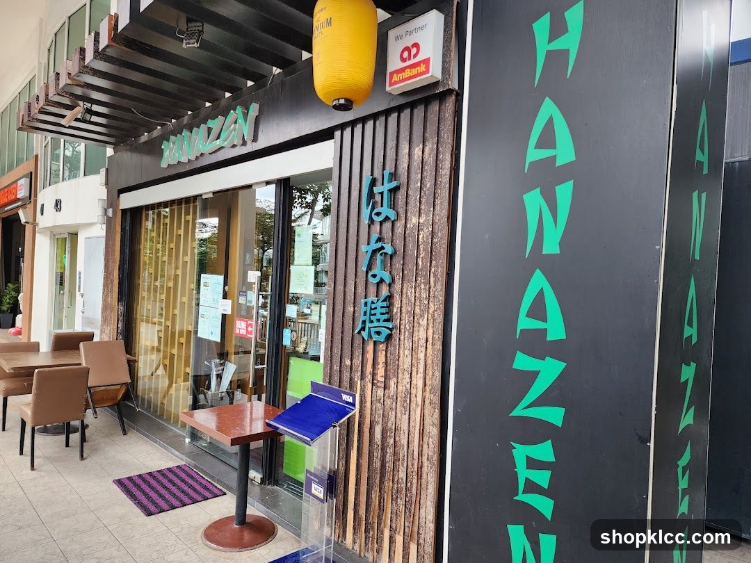 The exterior of a Japanese restaurant called Hanazen. The storefront features a wooden facade with vertical bamboo-like panels. The restaurant's name, "HANAZEN," is displayed in green lettering on a black vertical sign to the right of the entrance. Above the entrance, the name "HANAZEN" is also visible in green lettering. There are outdoor tables and chairs set up on the sidewalk in front of the restaurant. A yellow lantern hangs above the entrance.