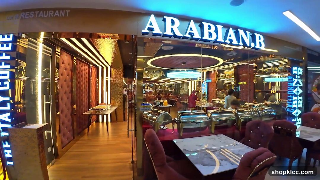 The exterior of a restaurant with a large sign that reads "ARABIAN.B" in illuminated white letters. To the left, a vertical neon sign reads "ITALY COFFEE". The entrance features glass doors and is flanked by plush, tufted red walls. Inside, a buffet setup with chafing dishes is visible, along with dining tables and chairs.