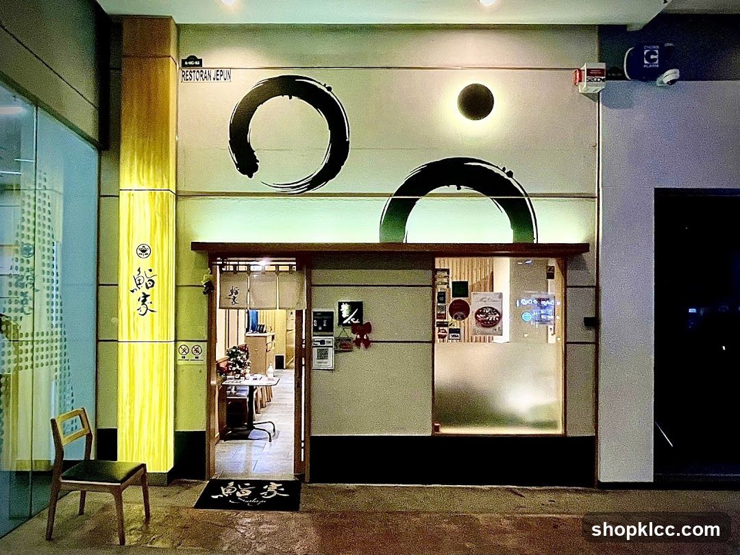 The image shows the exterior entrance of a restaurant, featuring large circular brushstroke designs on the facade and vertical Japanese characters on a wooden pillar. A glass door leads into the interior, where tables and chairs are visible. A welcome mat with Japanese text lies at the entrance.