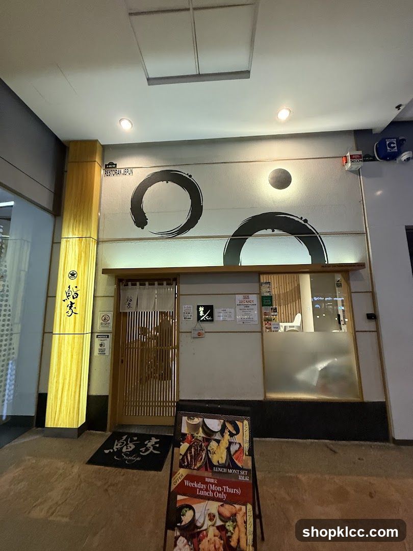 A Japanese restaurant entrance featuring a wooden gate with noren curtains, illuminated vertical signage with calligraphy, and large brushstroke circles on the wall above. A floor mat with matching calligraphy lies at the door. An A-frame menu board stands outside advertising lunch sets.