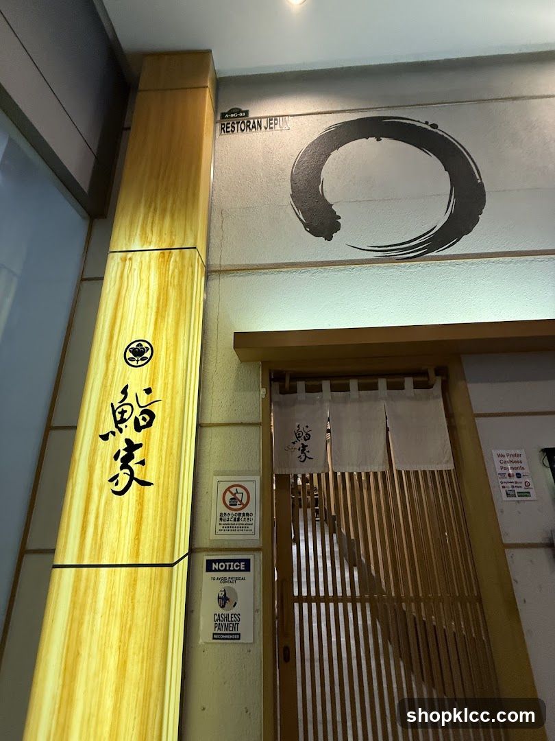 The image shows the entrance of a restaurant featuring traditional Japanese design elements. A wooden pillar displays vertical calligraphy in black ink, likely representing the establishment's name or theme. Above the entrance is a large circular brushstroke logo on a light-colored wall. The doorway is framed by bamboo slats and adorned with hanging white curtains bearing similar calligraphic characters. Several signs are visible: one prohibits smoking inside; another indicates cashless payment options; and a third mentions free Wi-Fi access. The overall aesthetic suggests an authentic or themed dining experience.