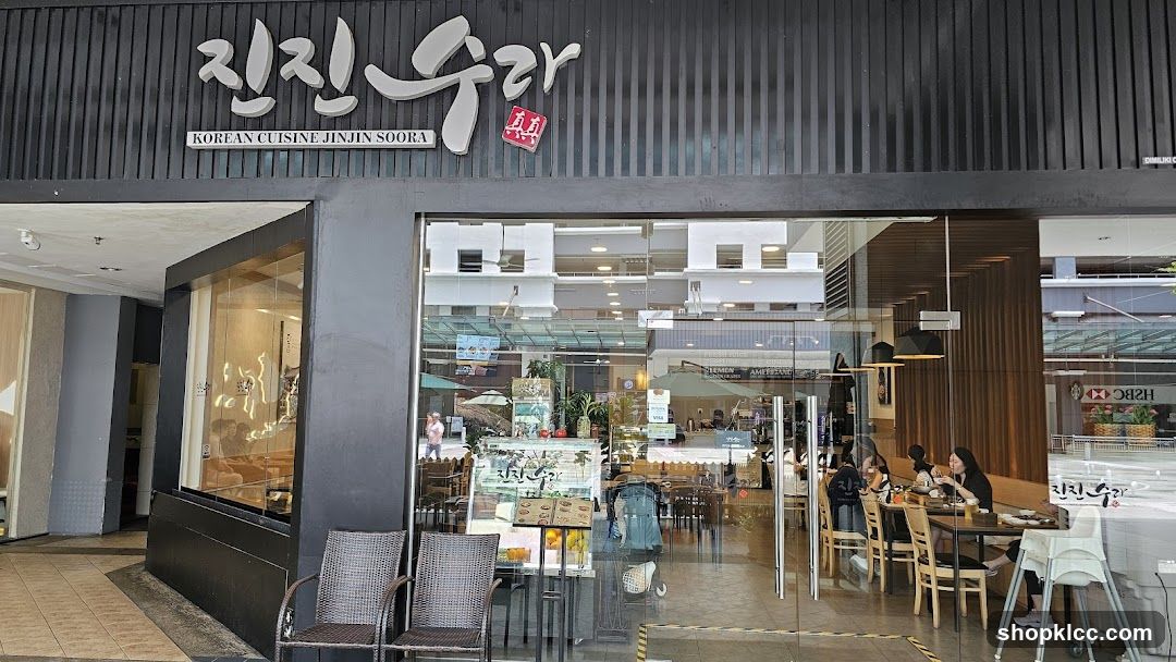 The image shows the storefront of Jinjin Soora, a Korean cuisine restaurant. The sign above the entrance displays '진진수라' in large stylized characters with 'KOREAN CUISINE JINJIN SOORA' written below it. The building facade is dark gray with vertical slats. Large glass windows and doors provide visibility into the interior dining area where patrons are seated at tables. A small display stand near the entrance holds brochures or menus. Two wicker chairs sit outside on the tiled walkway.