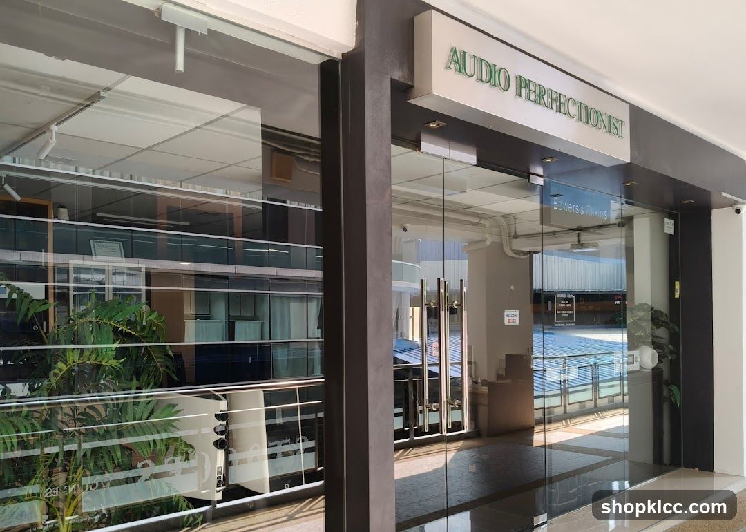 The exterior of a modern shop with large glass doors and windows. A prominent sign above the entrance reads "AUDIO PERFECTIONIST". Another sign inside the glass doors indicates "Bowers & Wilkins". A small "Welcome" sign is also visible. The reflection in the glass shows a multi-story building with many windows and some greenery.