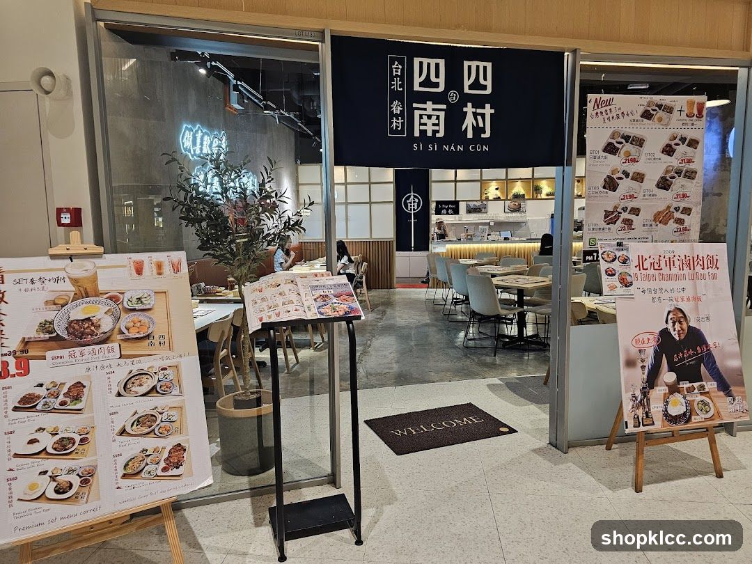 The exterior of a restaurant with a dark blue banner displaying the restaurant's name in Chinese and English. The entrance has large glass doors, and through them, tables and chairs are visible, along with a counter and kitchen area. Outside the entrance, there are several display boards with food images and prices, as well as a welcome mat.