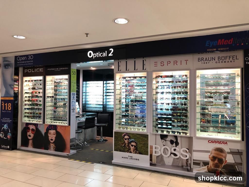The storefront of an optical shop named Optical 2. The shop displays various brands of eyeglasses and sunglasses in illuminated display cases. Brands visible include Police, Porsche Design, Gucci, Elle, Esprit, Braun Buffel, and Carrera. Promotional posters for Marc Jacobs and Boss are also displayed.
