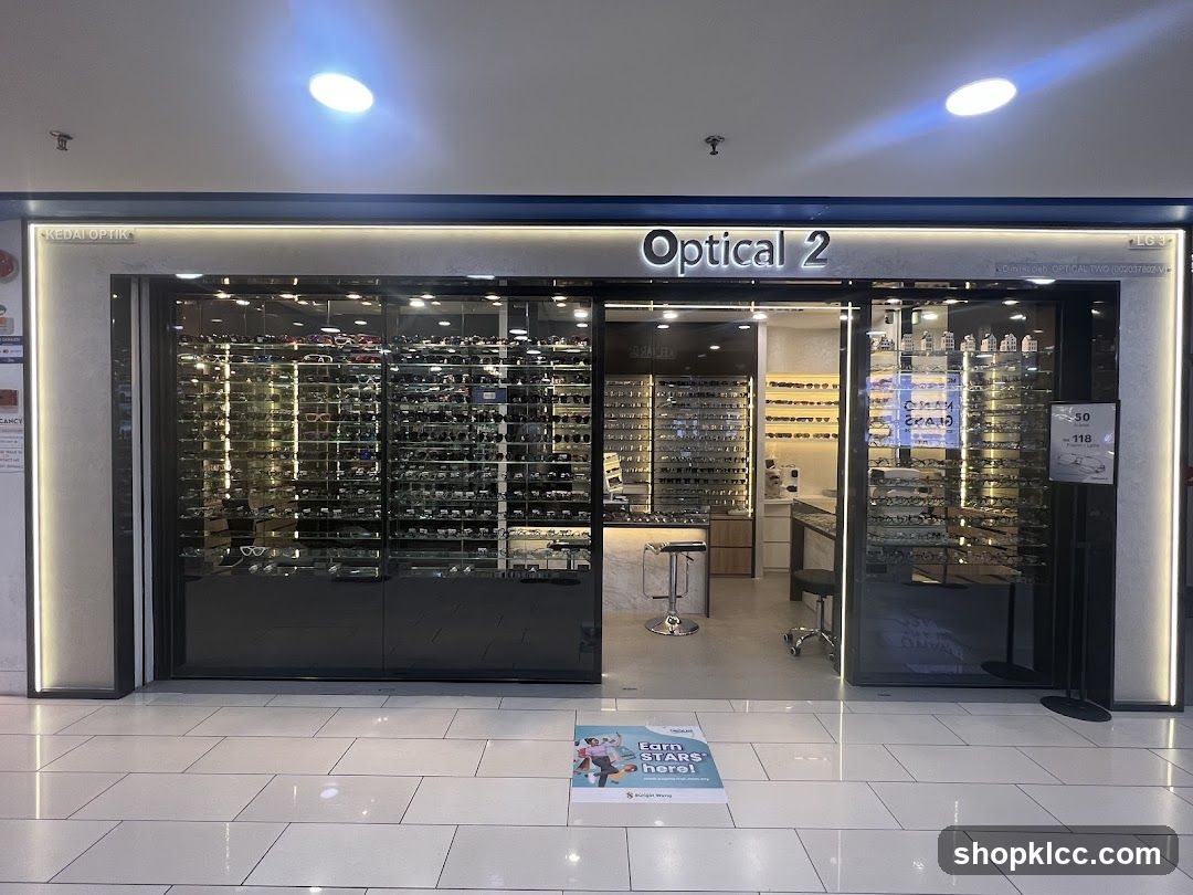 The exterior of an optical shop with large glass windows displaying numerous eyeglasses. The shop's name, "Optical 2", is prominently displayed above the entrance. The interior shows more eyewear displays and a counter area. A promotional sign is visible on the right side of the entrance.