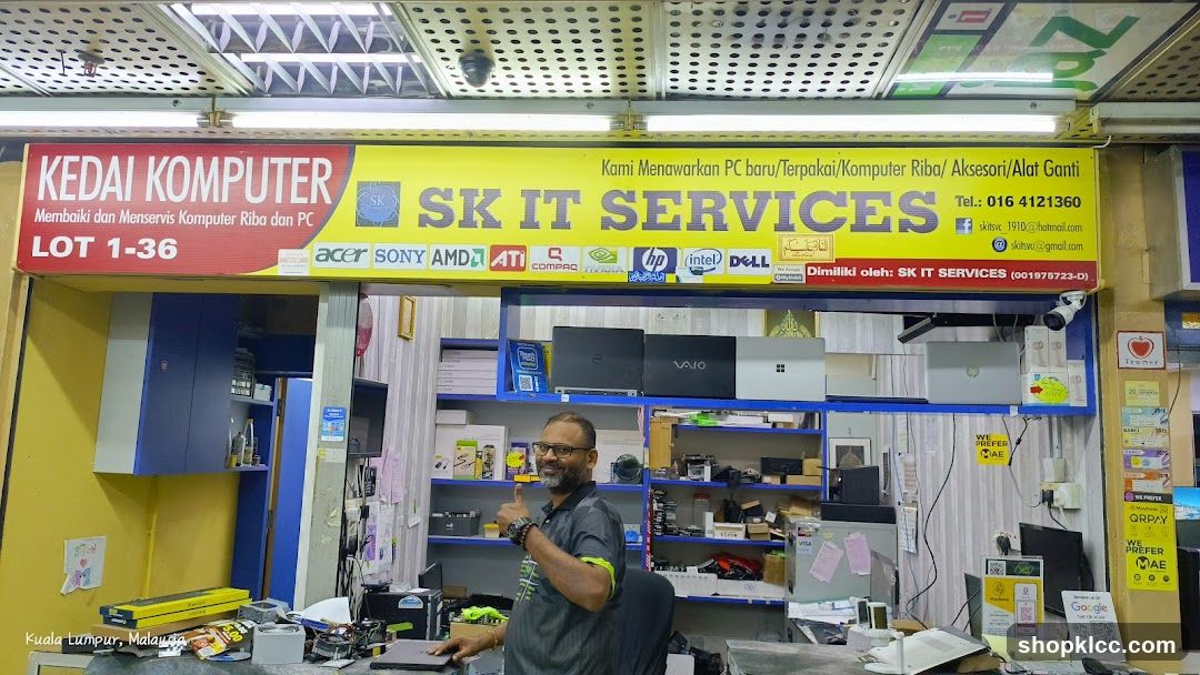 A computer shop with a yellow and red sign that says "KEDAI KOMPUTER" and "SK IT SERVICES". The sign also lists brands like Acer, Sony, AMD, ATI, Compaq, Nvidia, HP, Intel, and Dell. Below the sign, there are shelves with laptops and computer parts. A man is giving a thumbs up in front of the shelves. The shop is located in Kuala Lumpur, Malaysia.