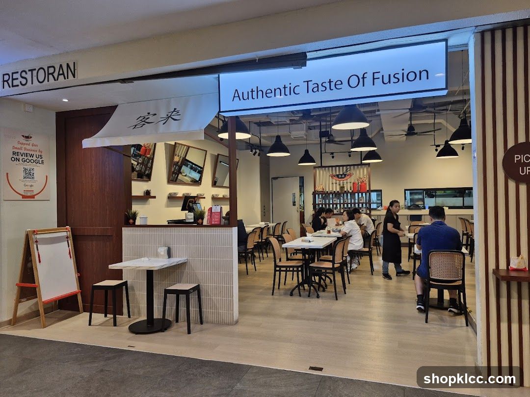 The exterior of a restaurant with a sign that reads "RESTORAN" and "Authentic Taste Of Fusion". There is a smaller sign with Chinese characters and a Google review QR code. Inside, tables and chairs are visible, with customers and staff present.