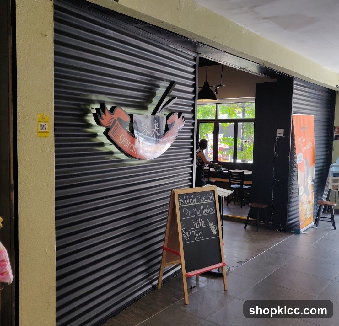 The image shows the exterior of a restaurant with a large, corrugated metal rolling door. A prominent illuminated sign with red and black lettering, featuring Chinese characters and the word "gerai.", is mounted on the door. To the left of the entrance, a yellow QR code is visible on the wall. In front of the entrance, a wooden A-frame chalkboard displays a daily special: "Sin-chu Meehun With Teh". Through the open doorway, tables and chairs are visible inside, along with a person standing near a window. To the right, a tall orange promotional banner is displayed.